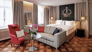 Een hotelkamer met een bed, een rode fauteuil, een grijze bank, een kleine tafel met een bloemenvaas, en een groot raam met gordijnen.