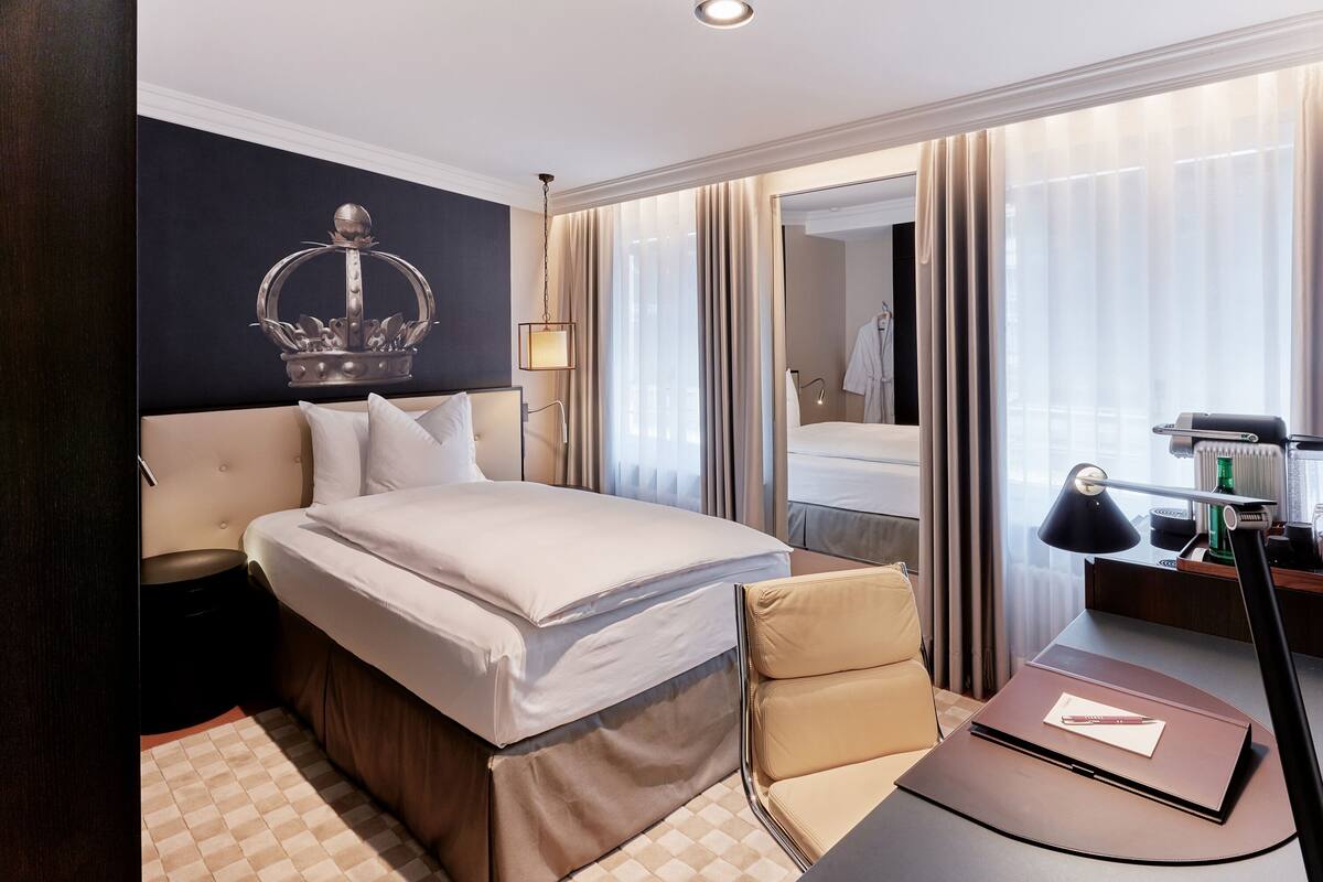 A hotel room with a large bed, a desk with a lamp, a mirror, and a window with curtains.