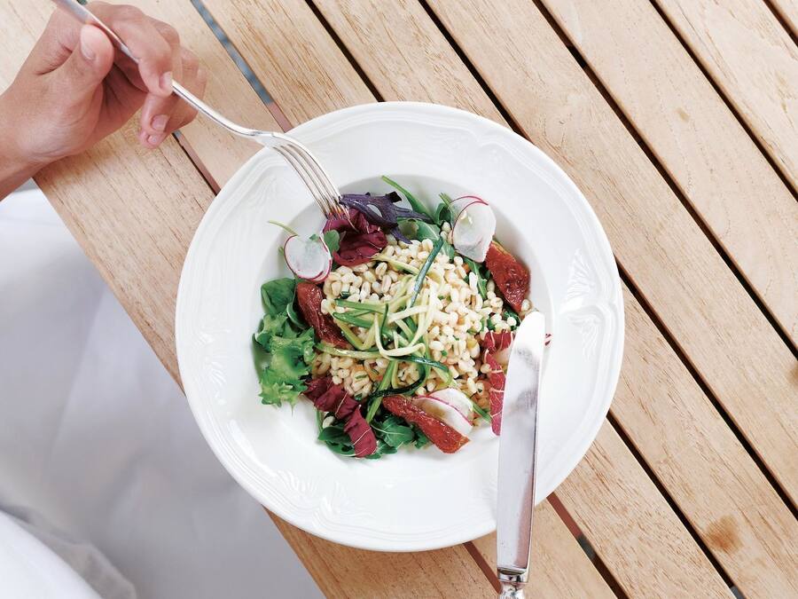 A wooden table with a white plate of salad, a fork, a knife, and a pair of sunglasses.