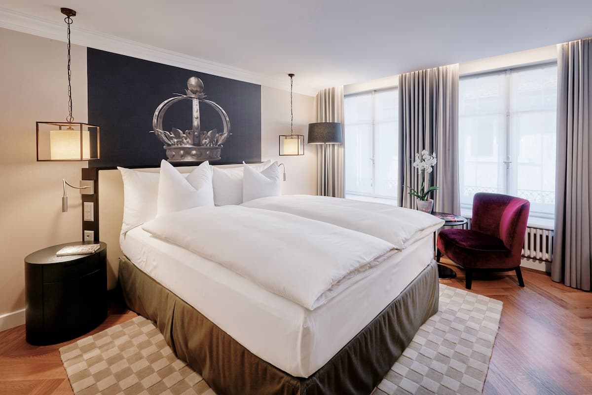 A large bed with white linens, a round nightstand, a small table with a lamp, and a chair.