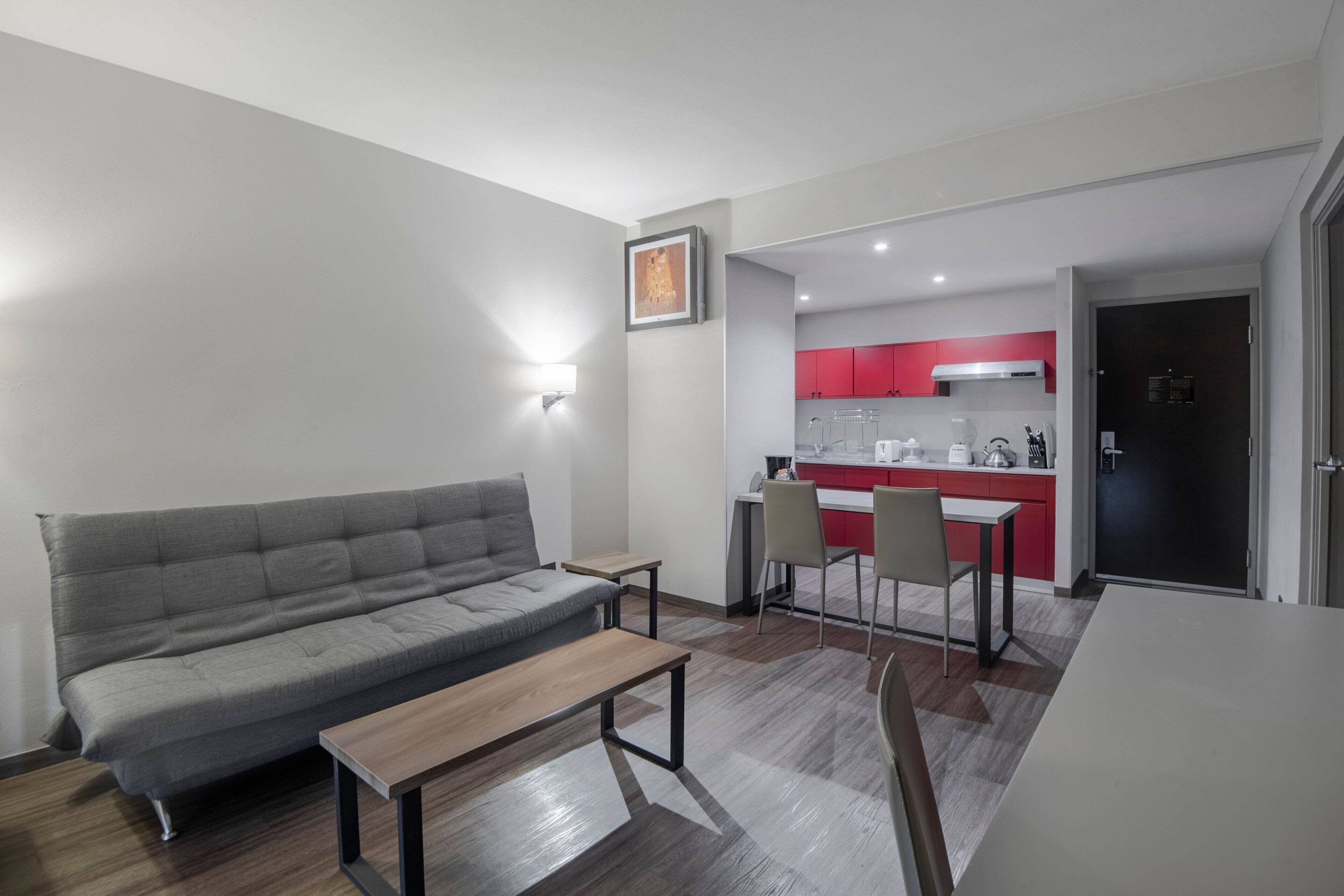 A modern living room with a grey sofa, a wooden coffee table, and a dining area with a white table and chairs. A kitchen with red cabinets and a black door is visible in the background.