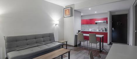 Un salon moderne avec un canapé gris, une table basse en bois et un coin repas comprenant une table et des chaises blanches. On aperçoit, au fond, une cuisine avec des placards rouges et une porte noire.