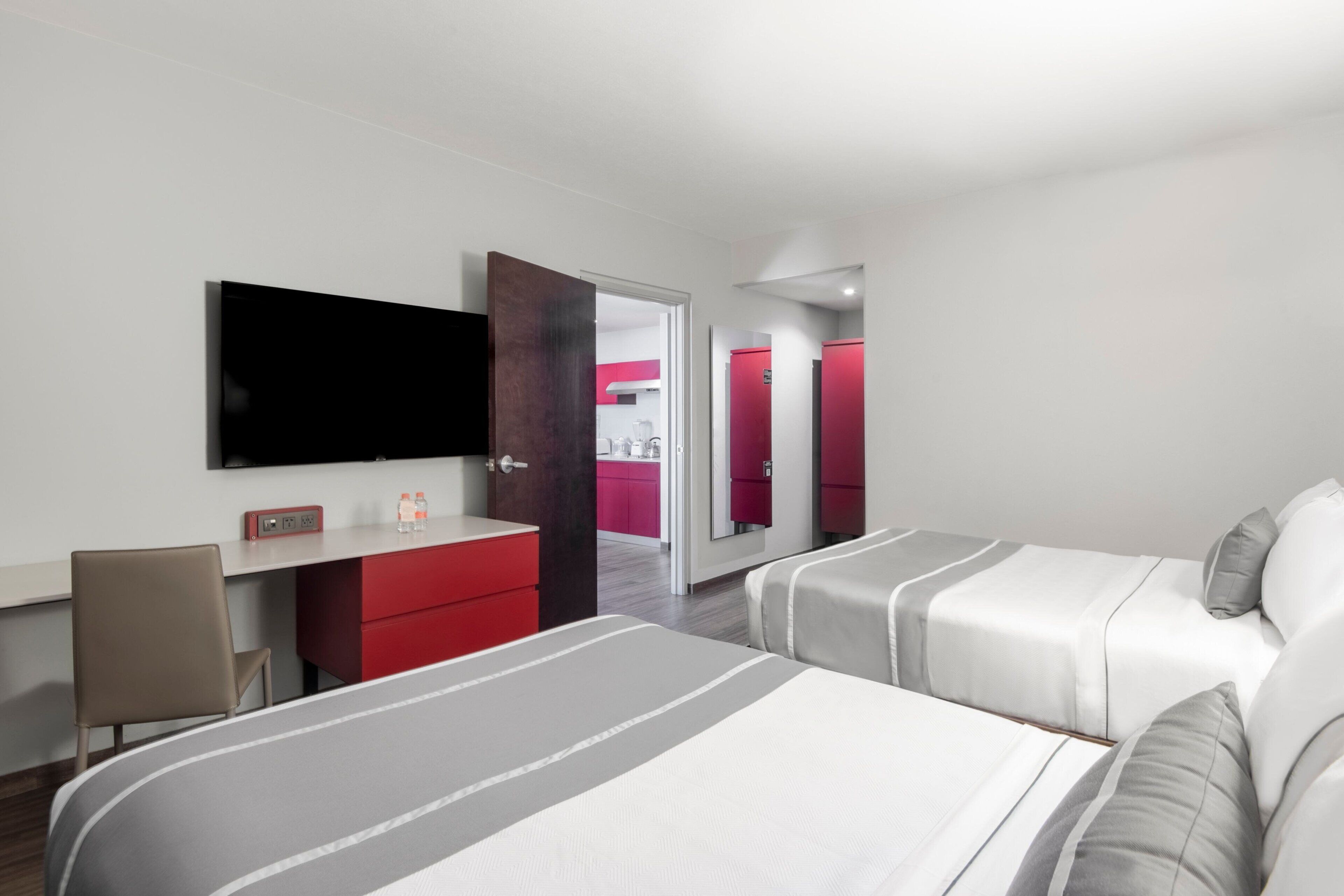 Une chambre d’hôtel moderne, équipée d’un lit, d’un bureau, d’une chaise, d’une télévision et d’une salle de bain visible à travers une porte ouverte.