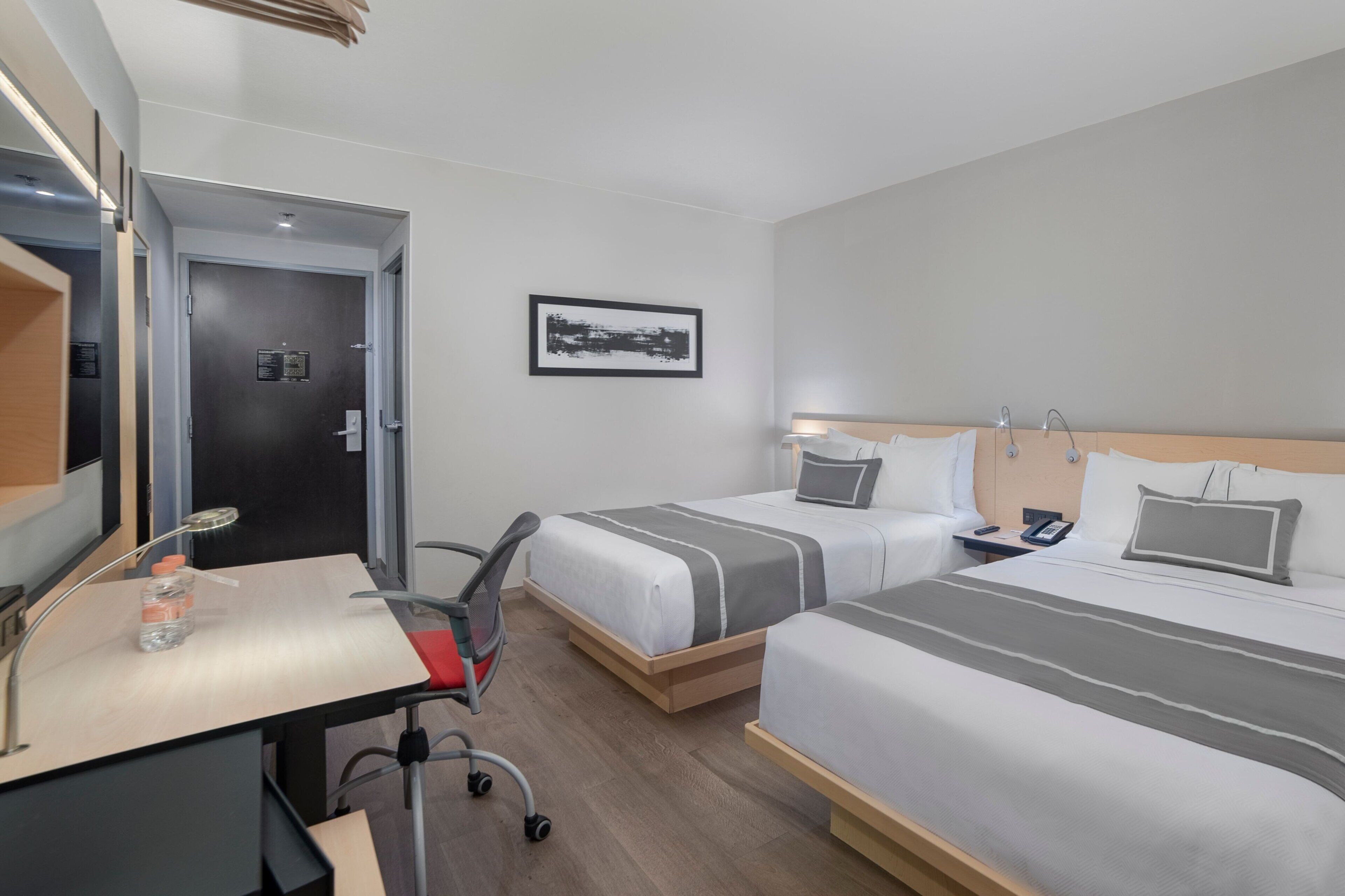 A hotel room with two beds, a desk, a chair, a small table, a mirror, and a framed picture on the wall.