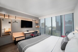 Ein modernes Hotelzimmer mit einem großen Bett, einem Schreibtisch, einem Sessel, einem Fernseher und einem großen Fenster mit Blick auf die Stadt.