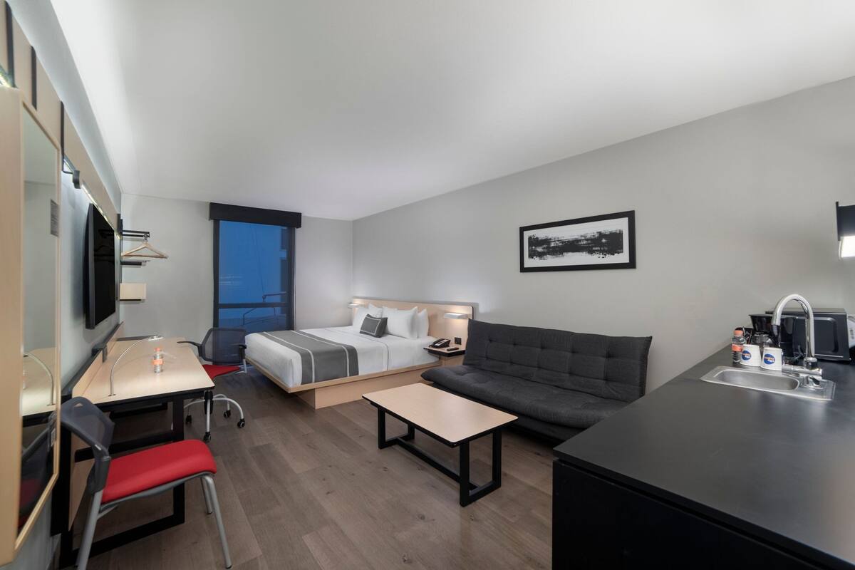 Moderni hotellihuone, jossa on sänky, sohva, työpöytä, tuoli, pieni pöytä, televisio, ikkuna, taulu seinällä ja pesuallas.