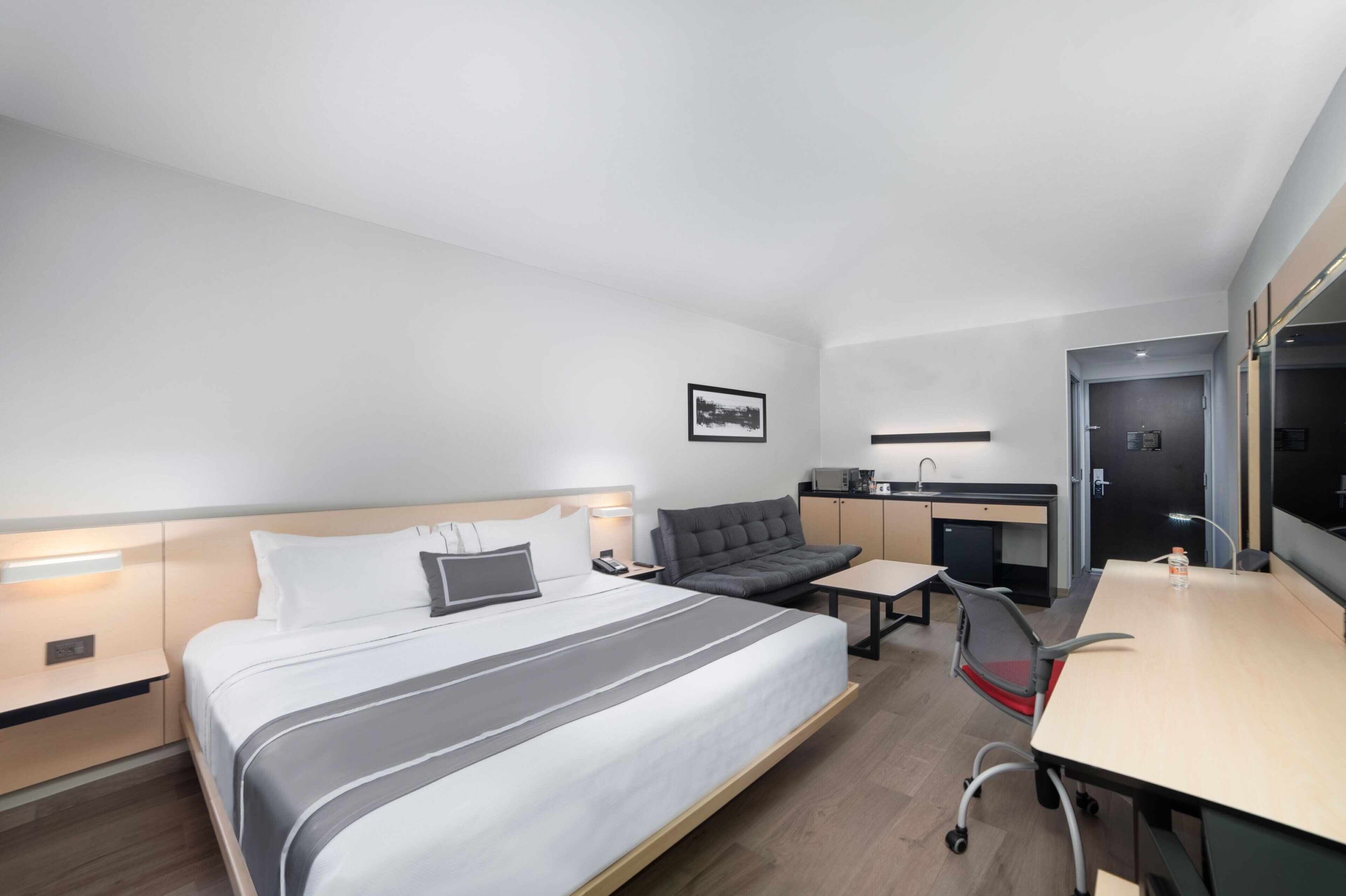 A modern hotel room with a large bed, a sofa, a desk, and a kitchenette.