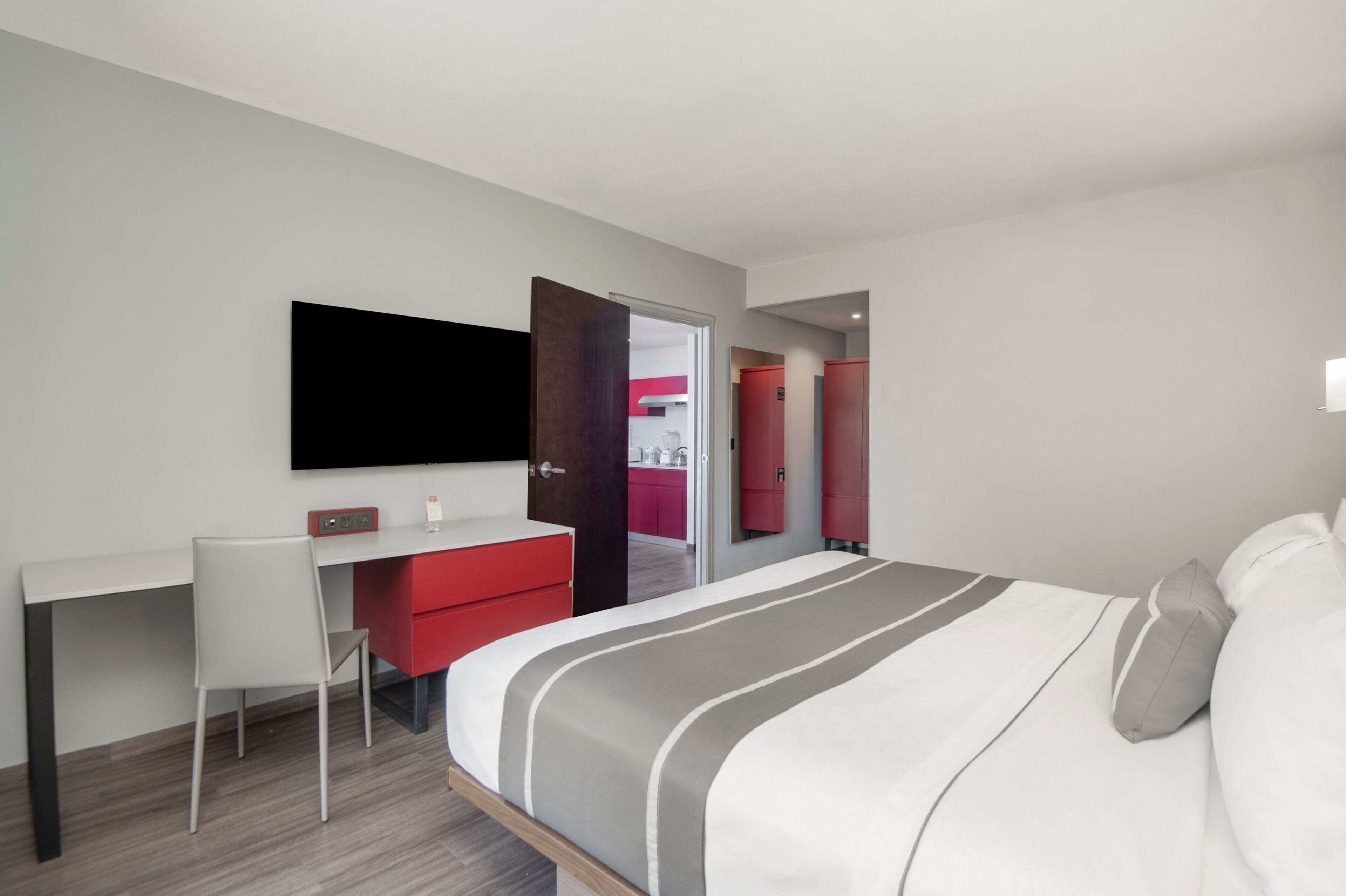 A modern hotel room with a bed, a desk, a chair, a TV, and a red wardrobe.