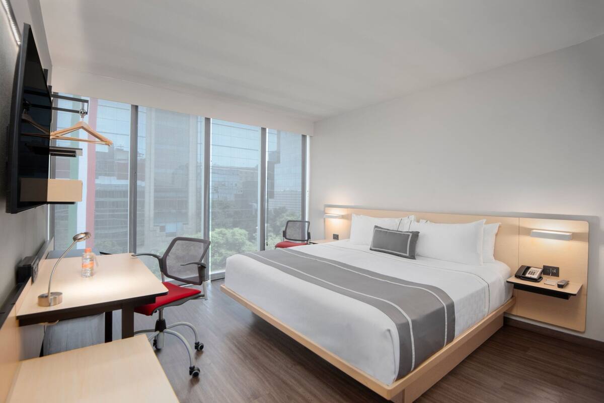 Moderni hotellihuone, jossa on suuri sänky, työpöytä tuolilla, seinälle asennettu televisio ja suuri ikkuna, josta avautuu näkymä kaupunkiin.