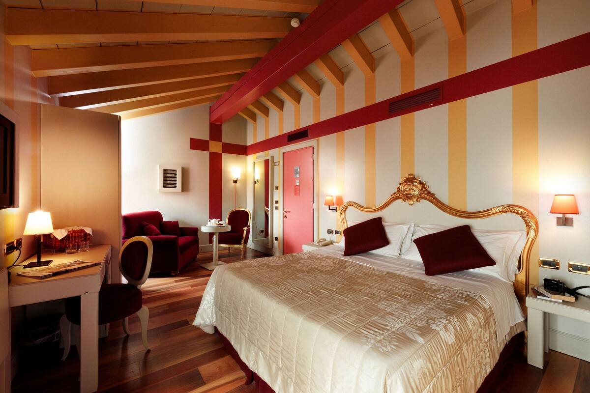 A hotel room with a large bed, a desk with a lamp, a chair, and a small table.