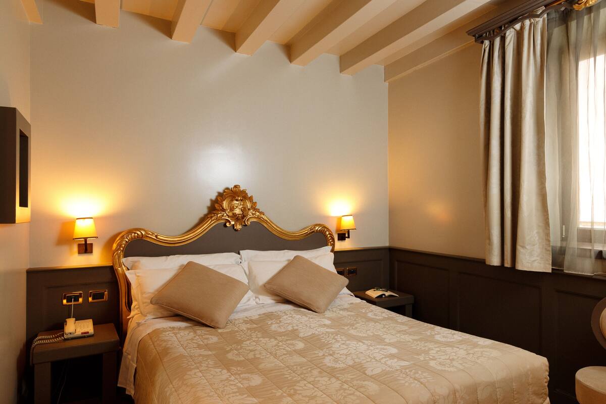 A hotel room with a large bed, two bedside lamps, a nightstand, and a window with curtains.