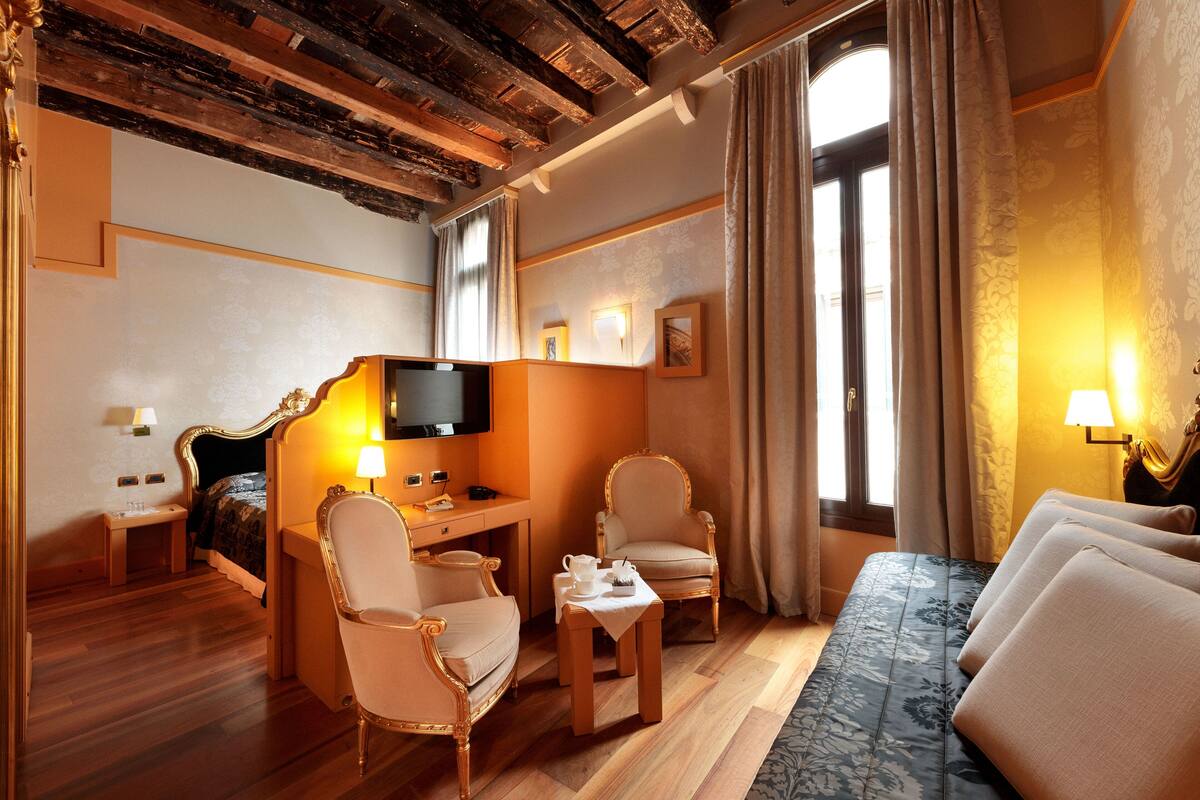 A hotel room with a bed, a desk with a computer, two armchairs, a small table with a lamp, and a TV on the wall.