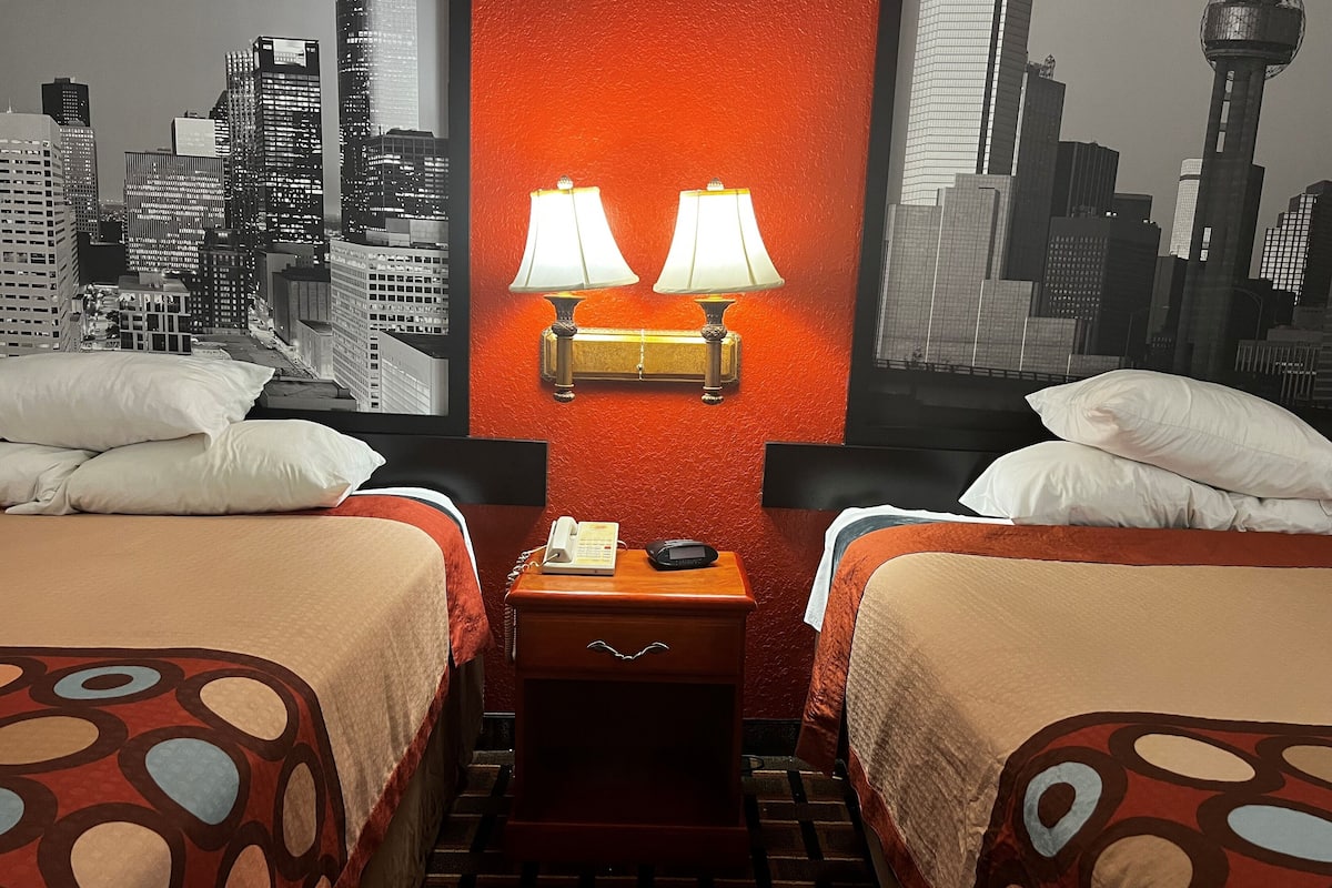 Hotelkamer met twee bedden, een nachtkastje met telefoon en lamp, en een muurschildering van een stadsgezicht.