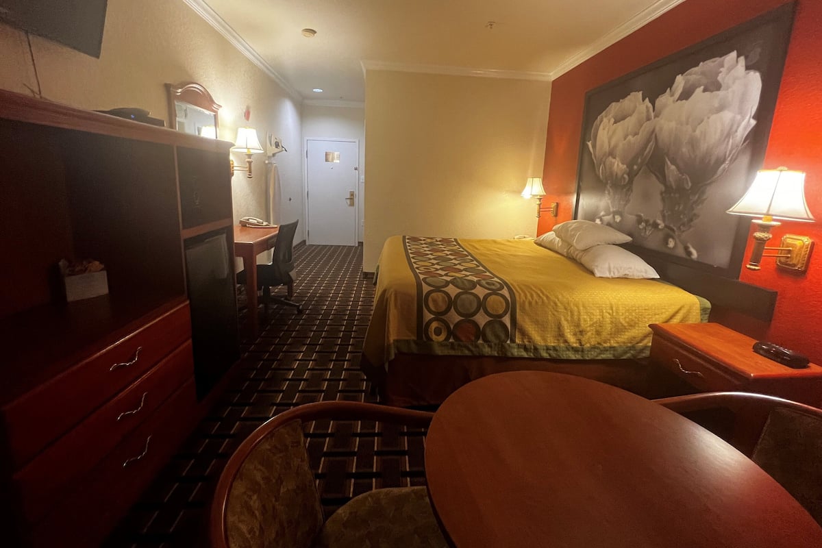 Een hotelkamer met een bed, een bureau met stoel, een klein tafeltje, een spiegel en een deur naar een gang.