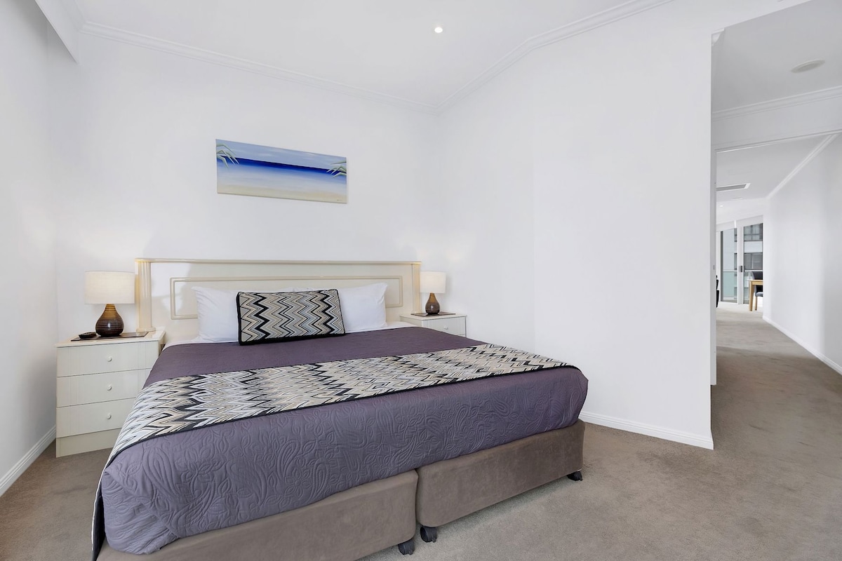 A bedroom with a bed, nightstands, a lamp, a painting, and a carpeted floor.