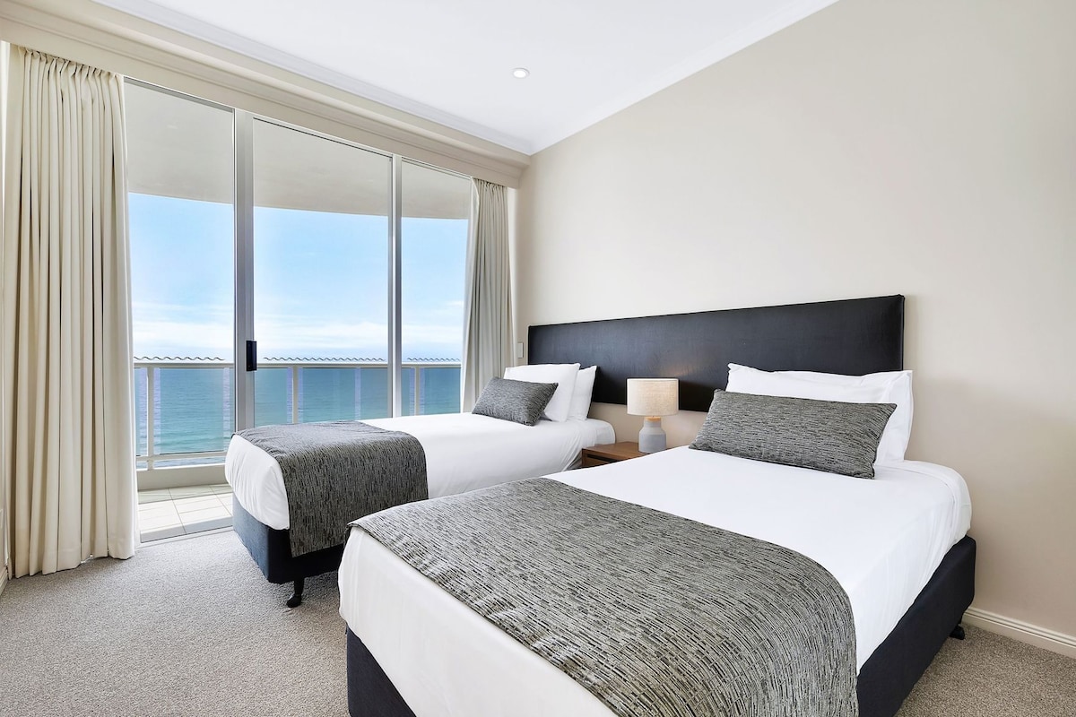 A hotel room with two beds, a nightstand, a lamp, and a view of the sea.