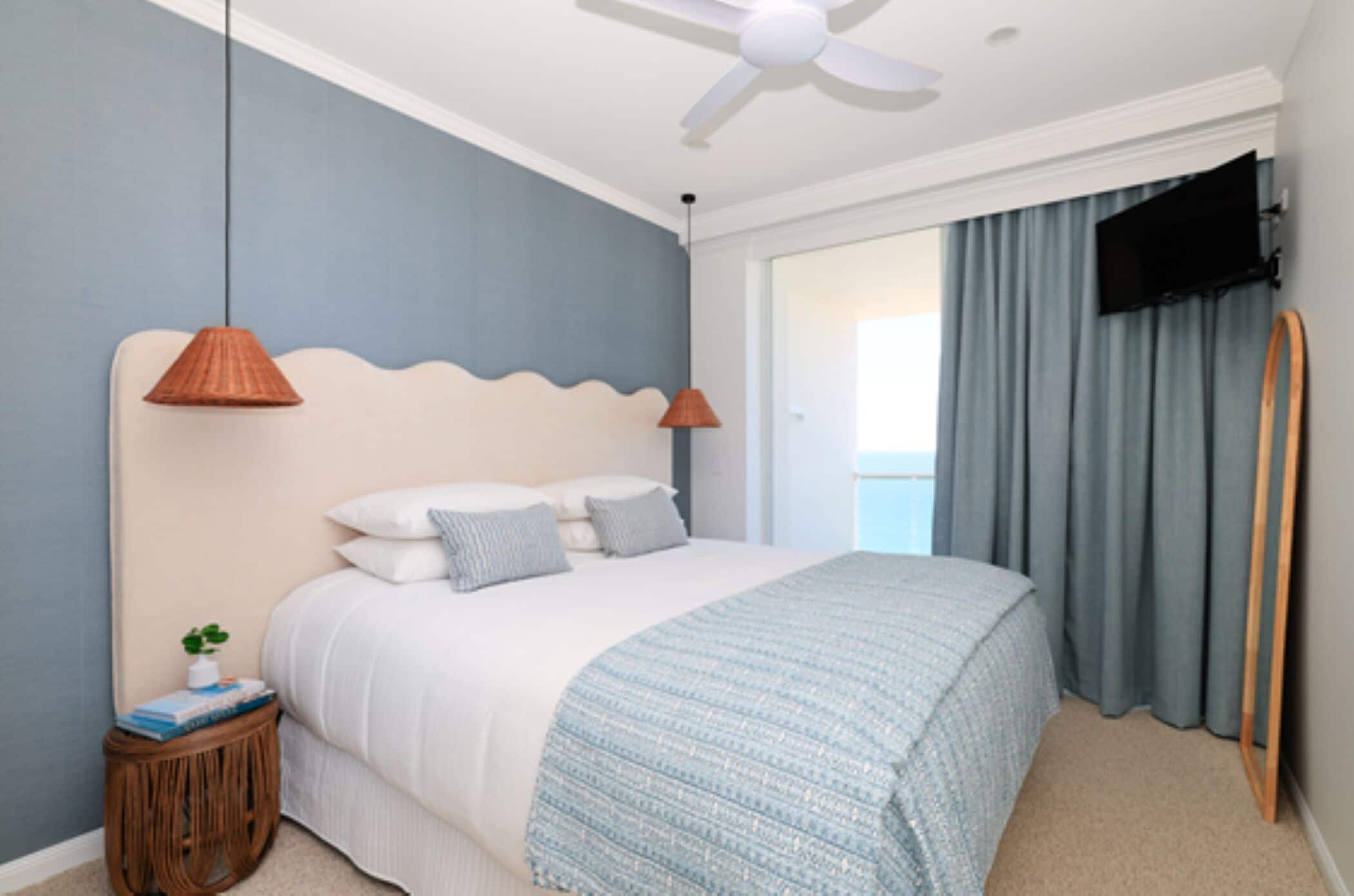 A bedroom with a bed, two pendant lights, a ceiling fan, a TV mounted on the wall, and a window with curtains.