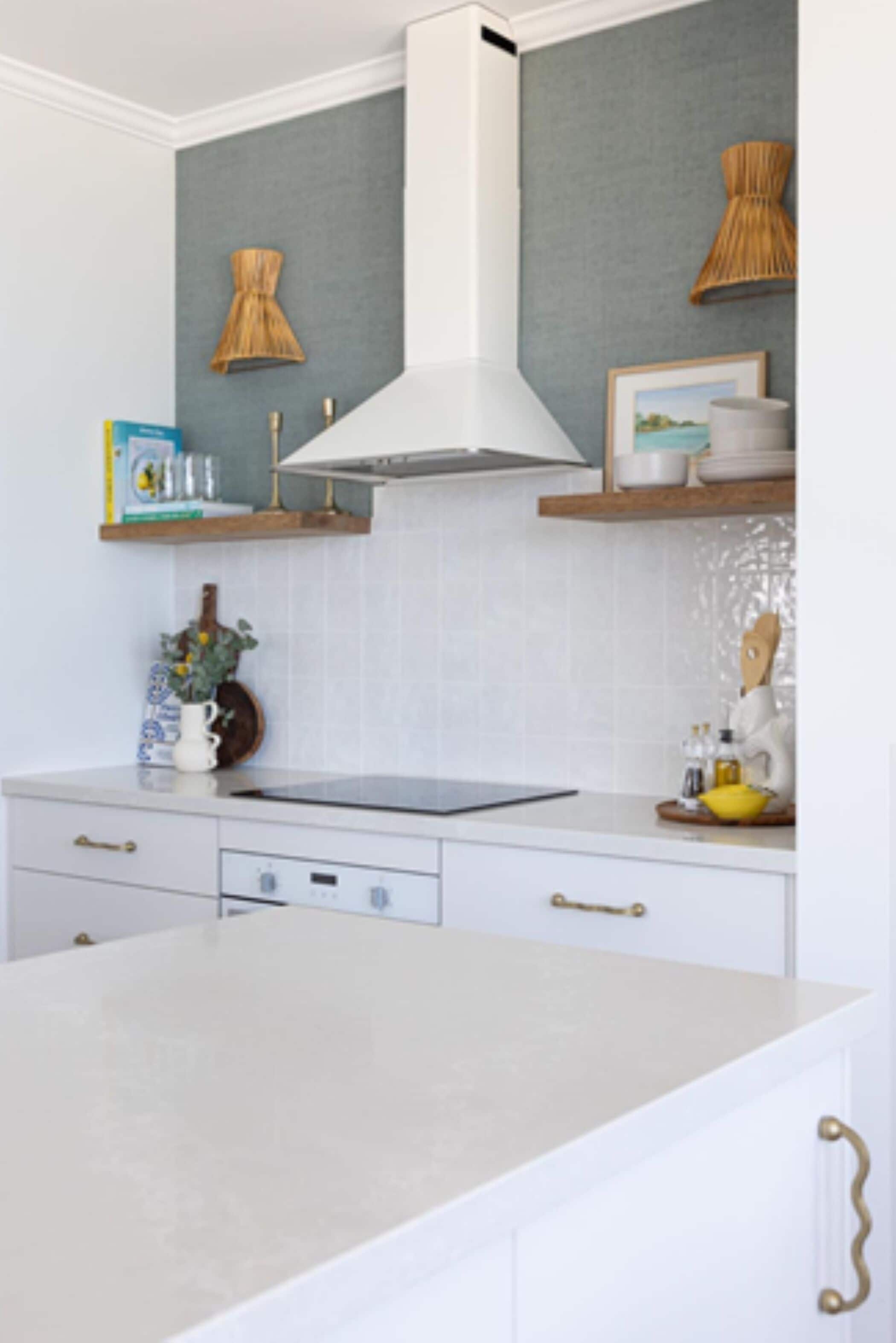 Una cocina con armarios blancos, una encimera blanca, una estufa empotrada y una campana extractora. Hay estantes de madera sobre la estufa con platos y una fotografía enmarcada.