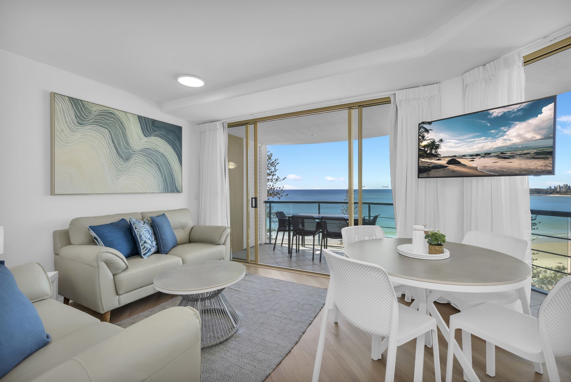 A modern living room with a sofa, coffee table, and dining area, featuring a large window with a view of the sea.