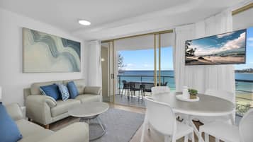 A modern living room with a sofa, coffee table, and dining area, featuring a large window with a view of the sea.