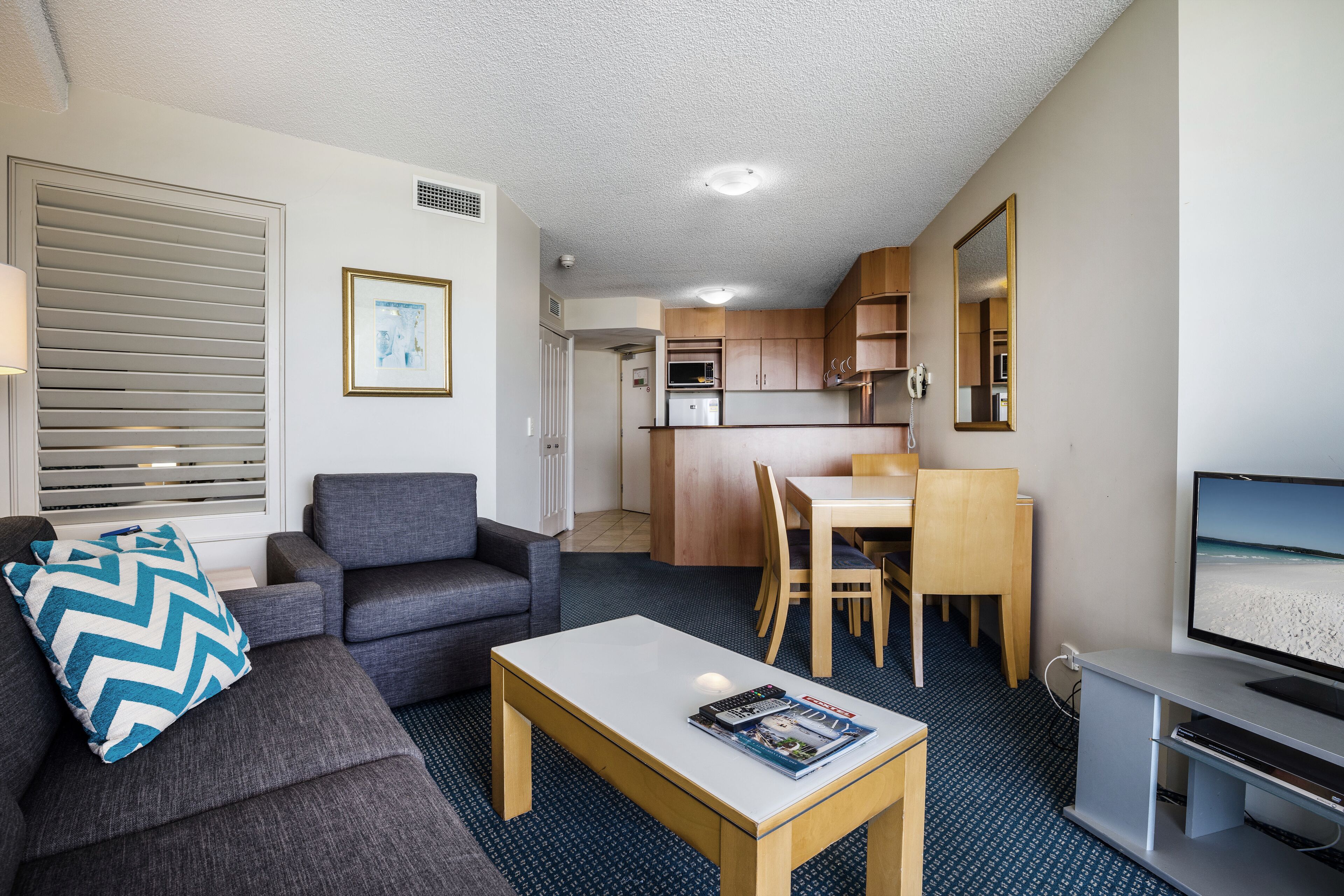A compact living space with a sofa, coffee table, and a television. A kitchenette is visible in the background.