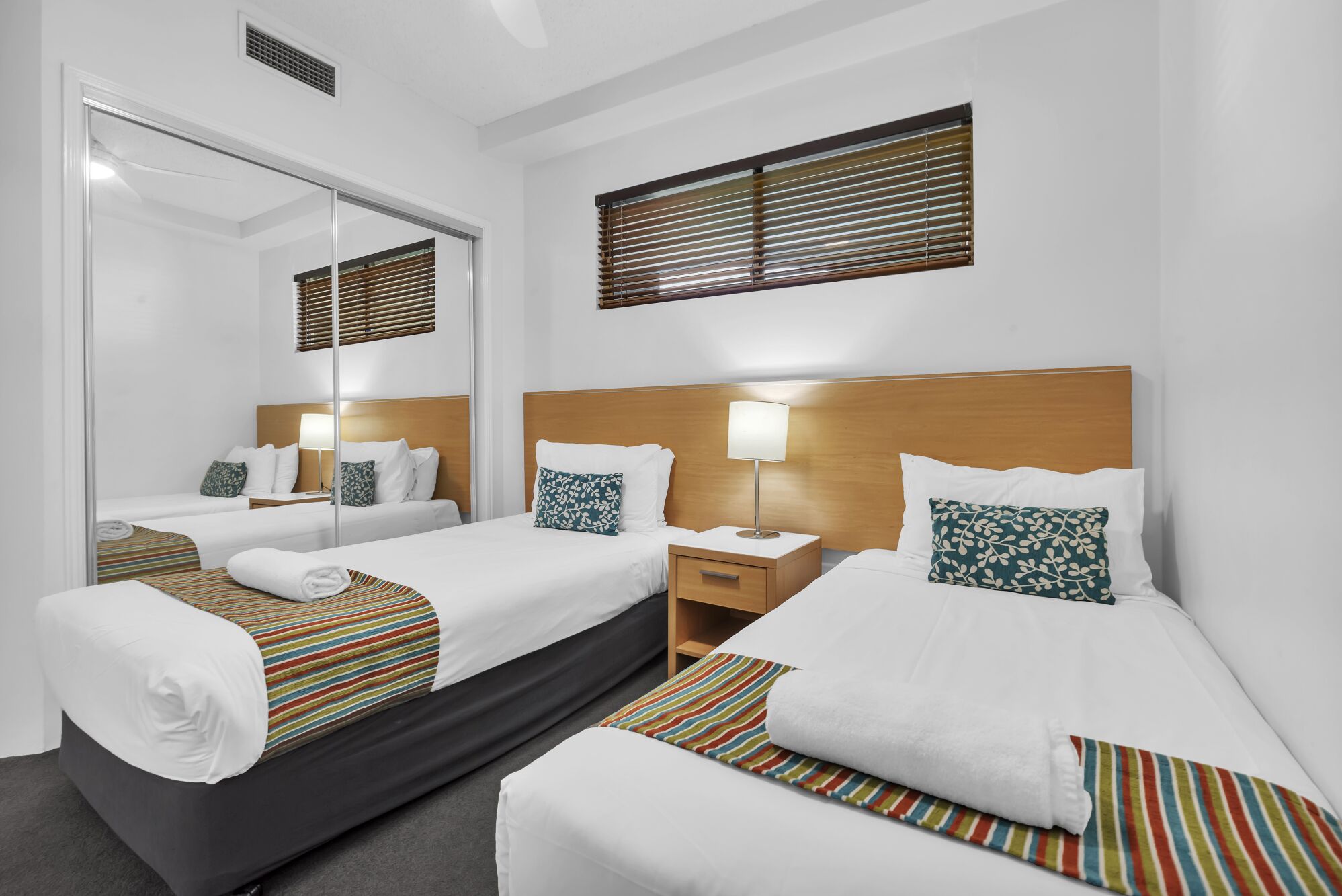A hotel room with two beds, a mirror, a nightstand with a lamp, and a window with blinds.