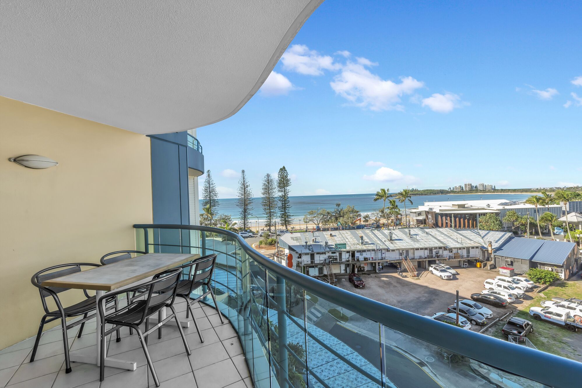 Un balcon avec une table et des chaises, donnant sur une plage et un parking où sont garées plusieurs voitures.