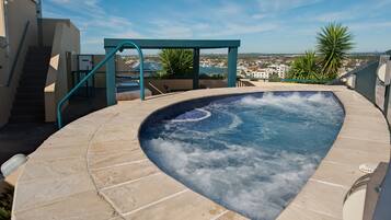 A hot tub with a curved edge, surrounded by beige tiles, with a view of a cityscape in the background.