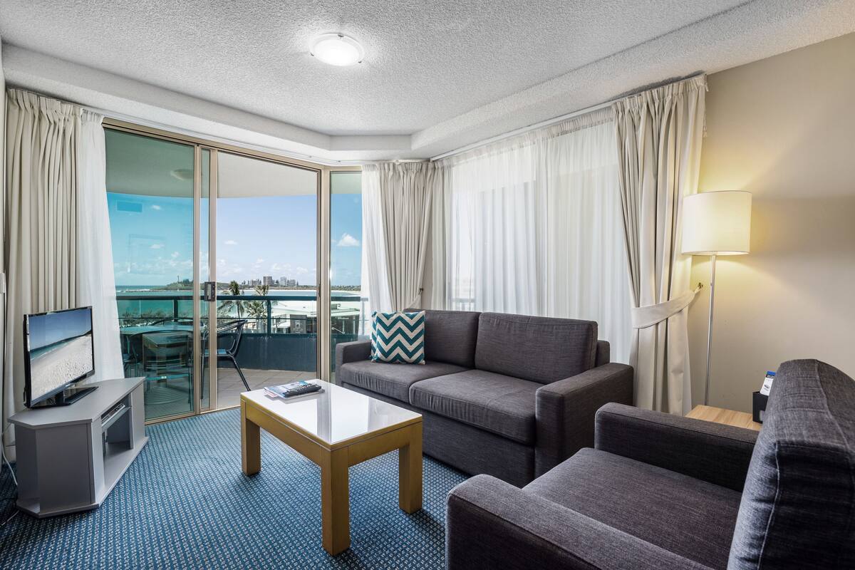 A modern hotel room with a sofa, a coffee table, a TV, and a balcony with a city view.