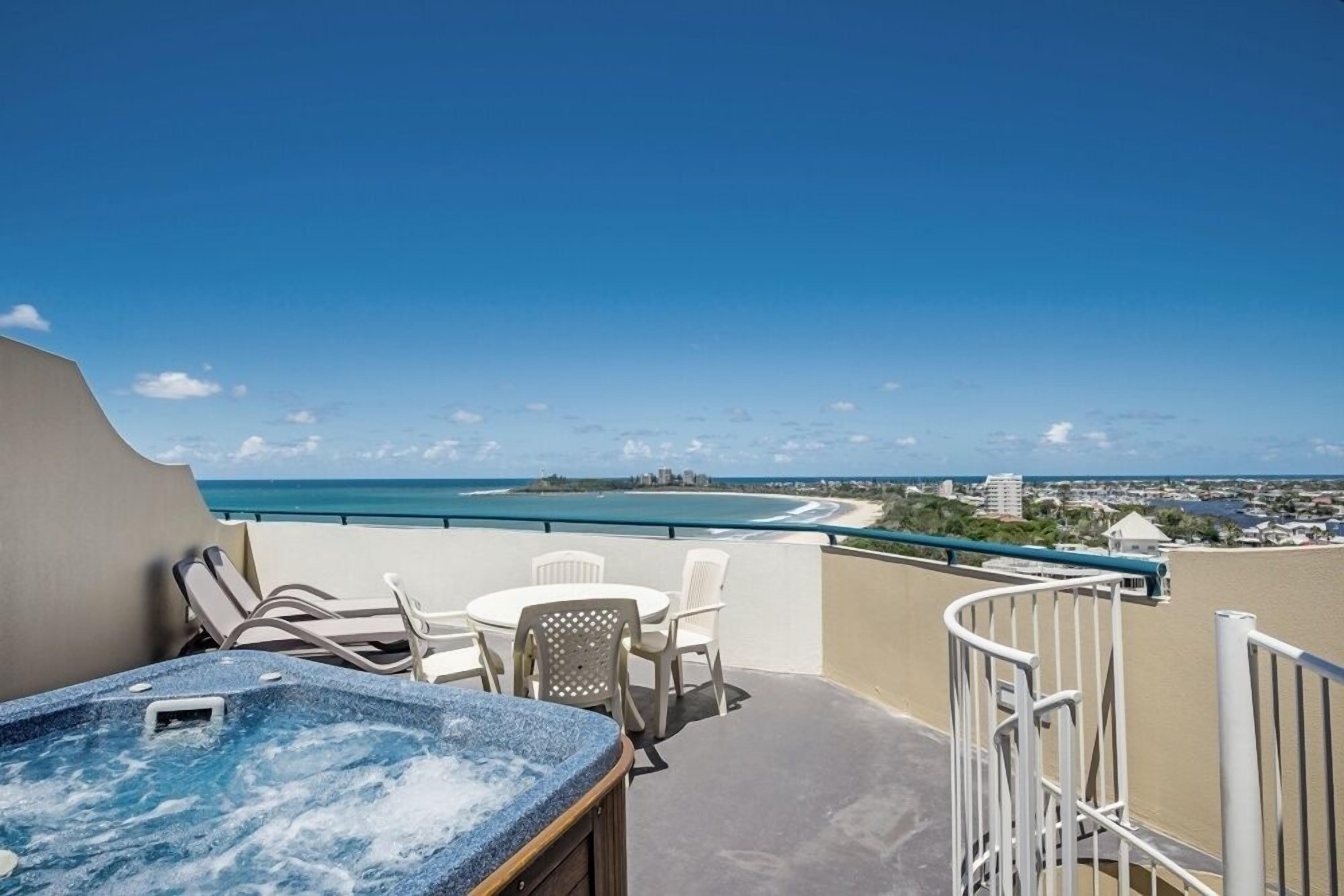 A balcony with a hot tub, white chairs, and a view of the beach and city skyline.