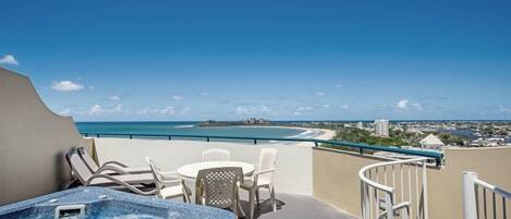 A balcony with a hot tub, white chairs, and a view of the beach and city skyline.