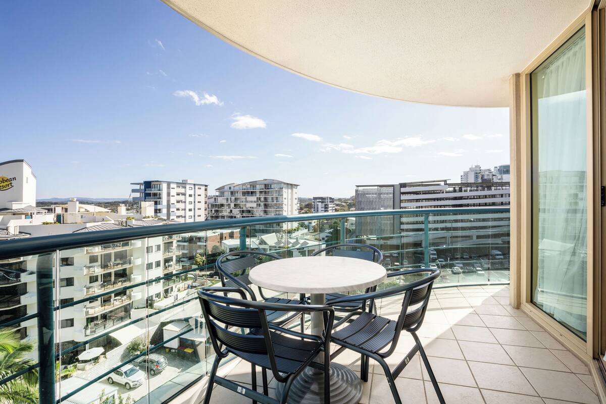 A balcony with a round table and chairs, overlooking a cityscape and a parking garage.