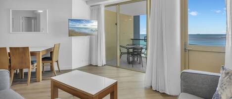 A modern living room with a dining area, a balcony with a view of the sea, and a sliding glass door leading to an outdoor seating area.