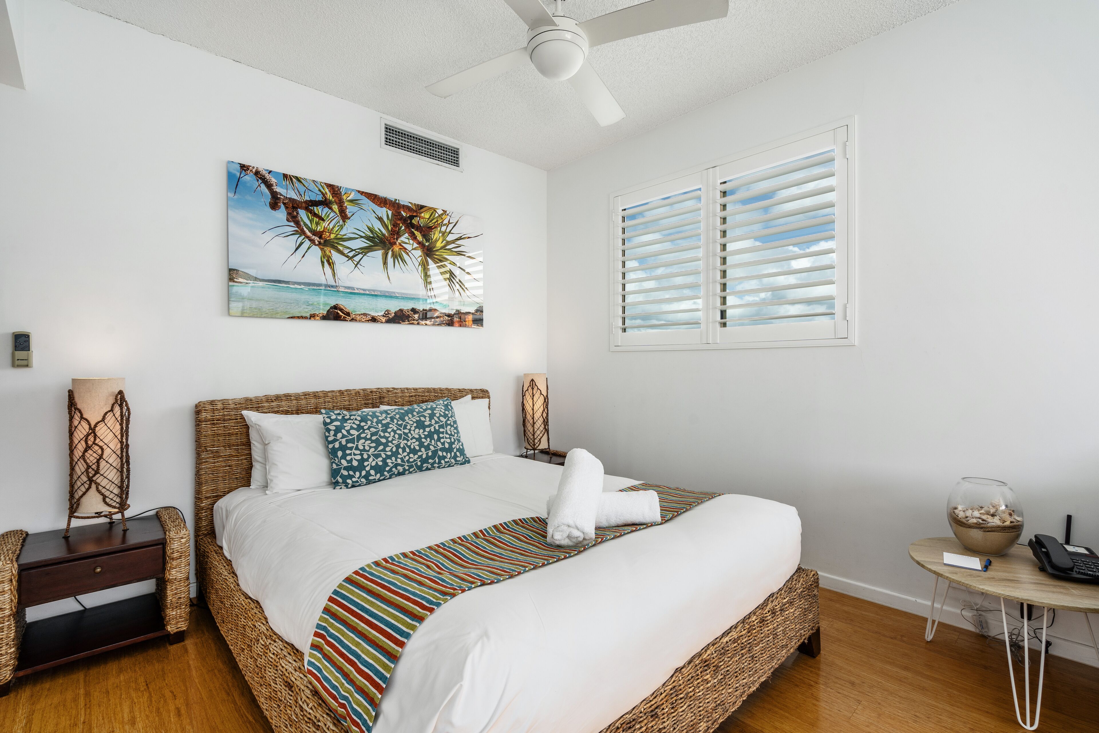 A neatly made bed with a striped bedspread, a tropical-themed painting above it, and a window with blinds.