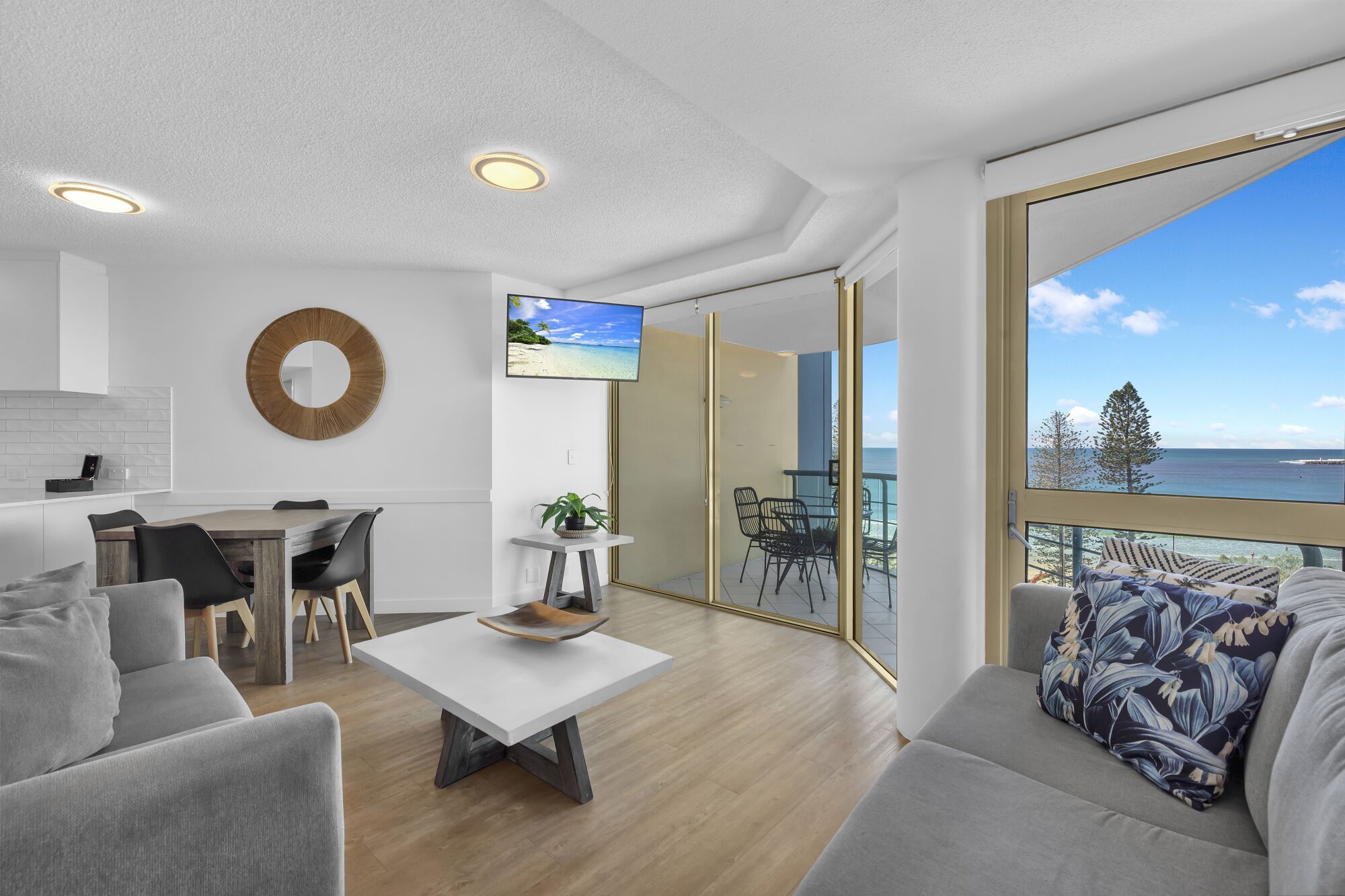 A modern living room with a dining area, a large window with a view of the ocean, and a balcony with outdoor furniture.