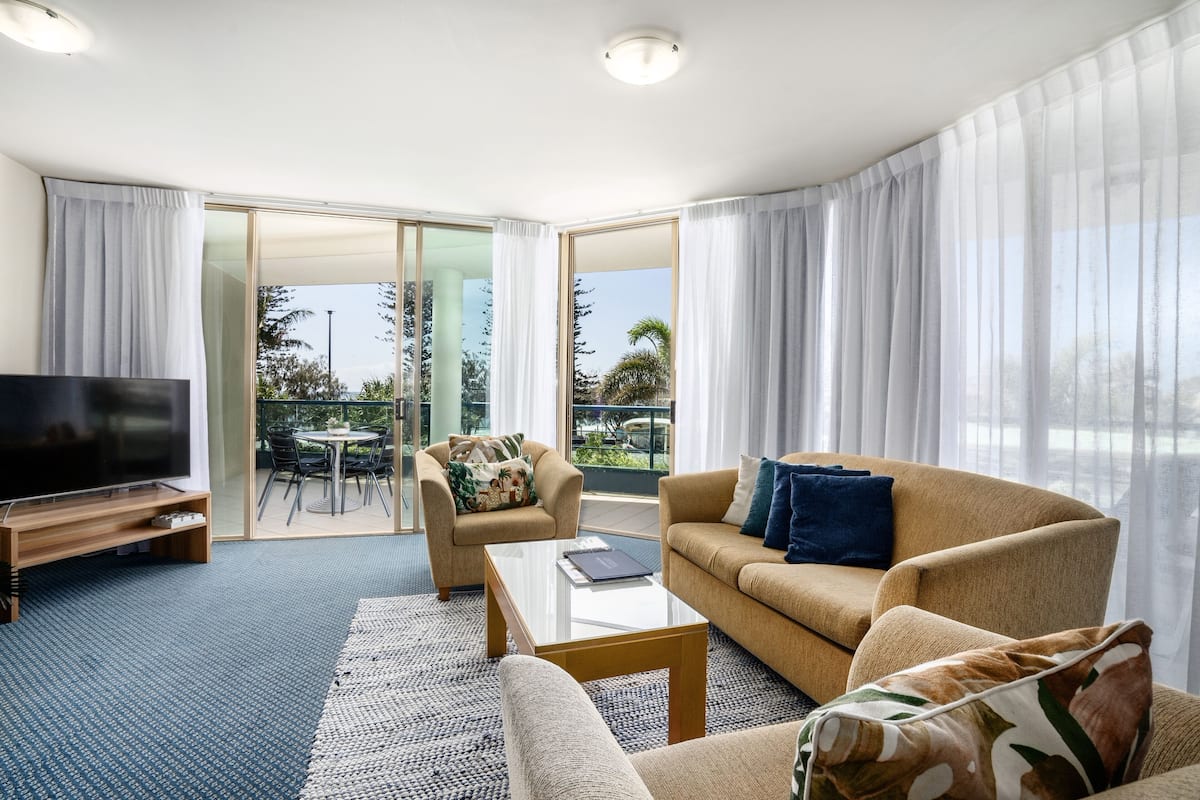 A modern living room with a sofa, armchair, and coffee table. There is a TV on a stand and a balcony with outdoor furniture.