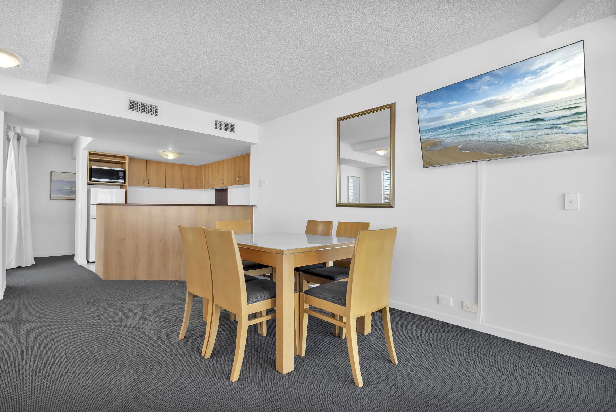 Ein kompaktes Hotelzimmer mit einer Küchenzeile, einem Essbereich mit Tisch und Stühlen, einem großen Spiegel und einem Gemälde mit Strandblick.