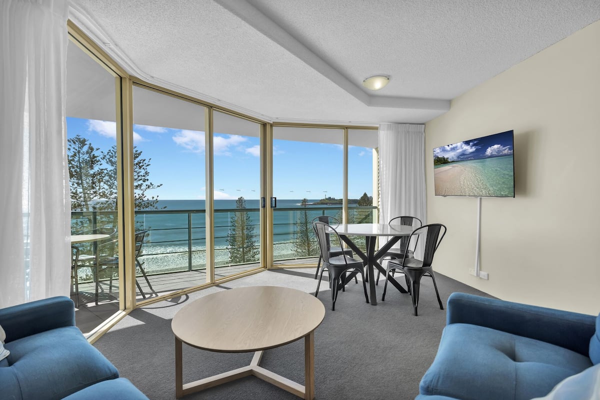 A room with a balcony, a dining table, chairs, a sofa, and a TV on the wall.