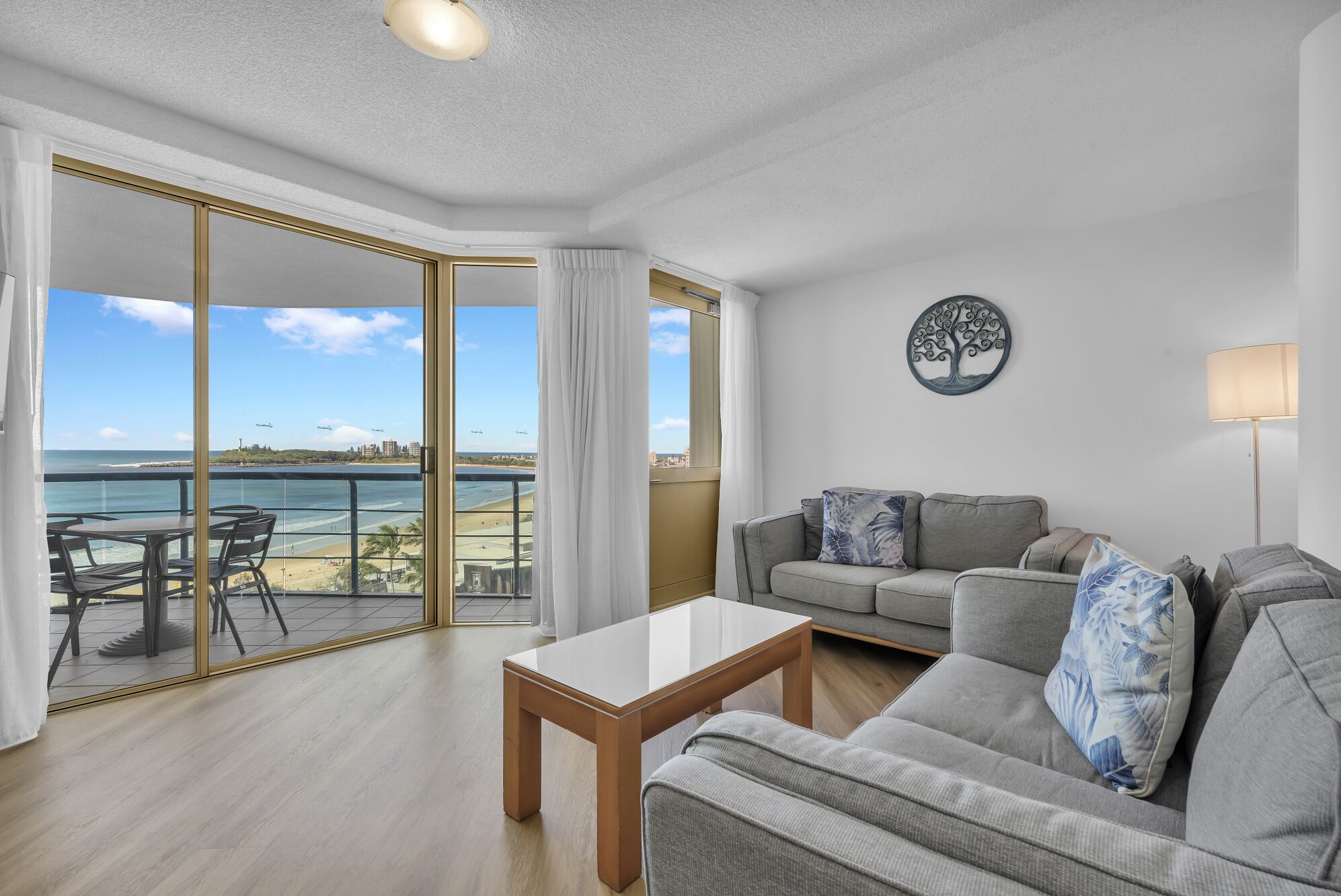A modern living room with a sofa, a coffee table, and a view of the beach through large glass doors.