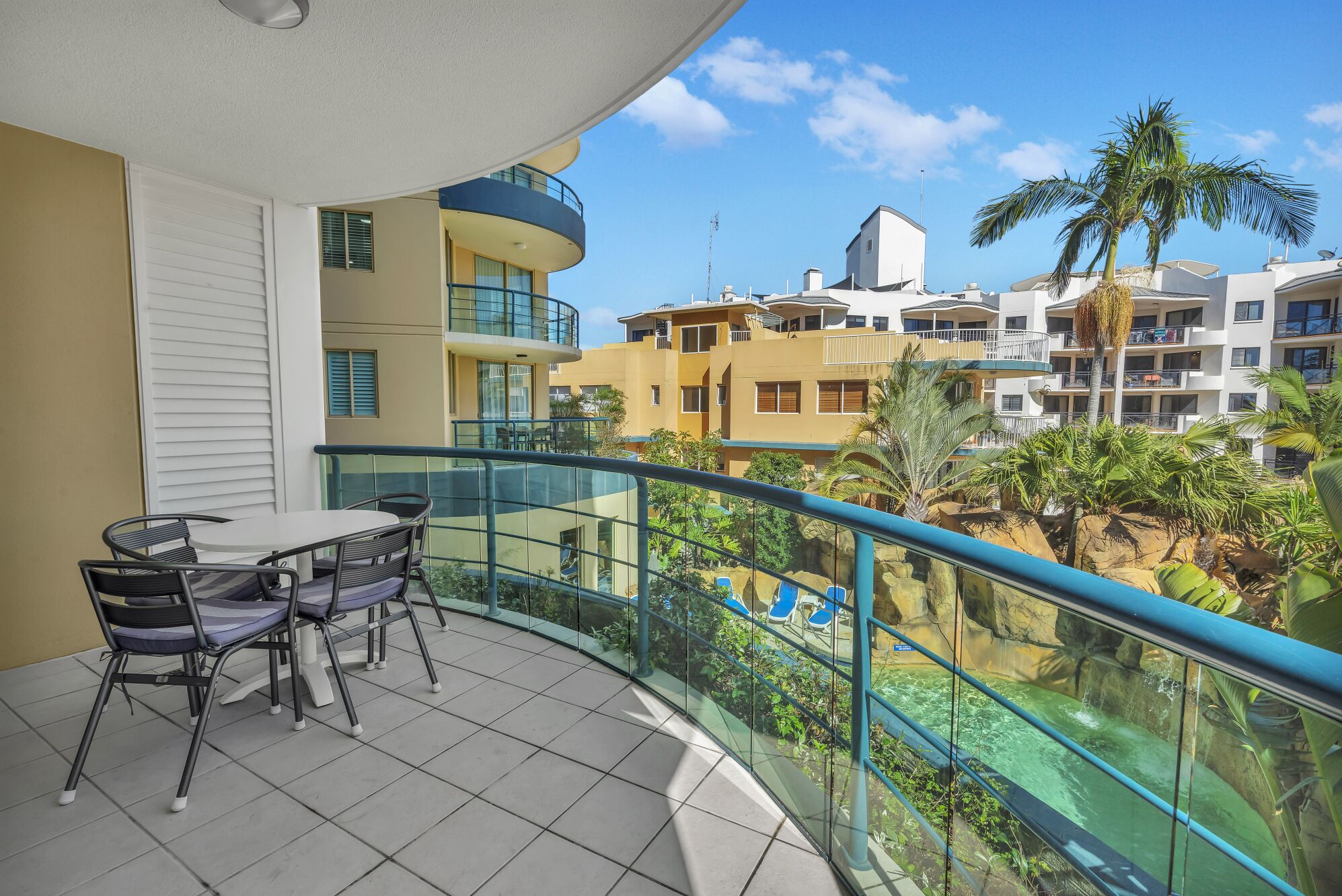 Un balcón con mesa y sillas, barandilla de cristal y vistas a una piscina y edificios.