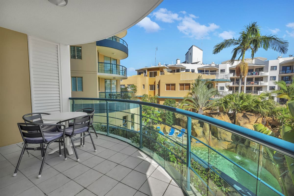 A balcony with a table and chairs, a glass railing, and a view of a pool and buildings.