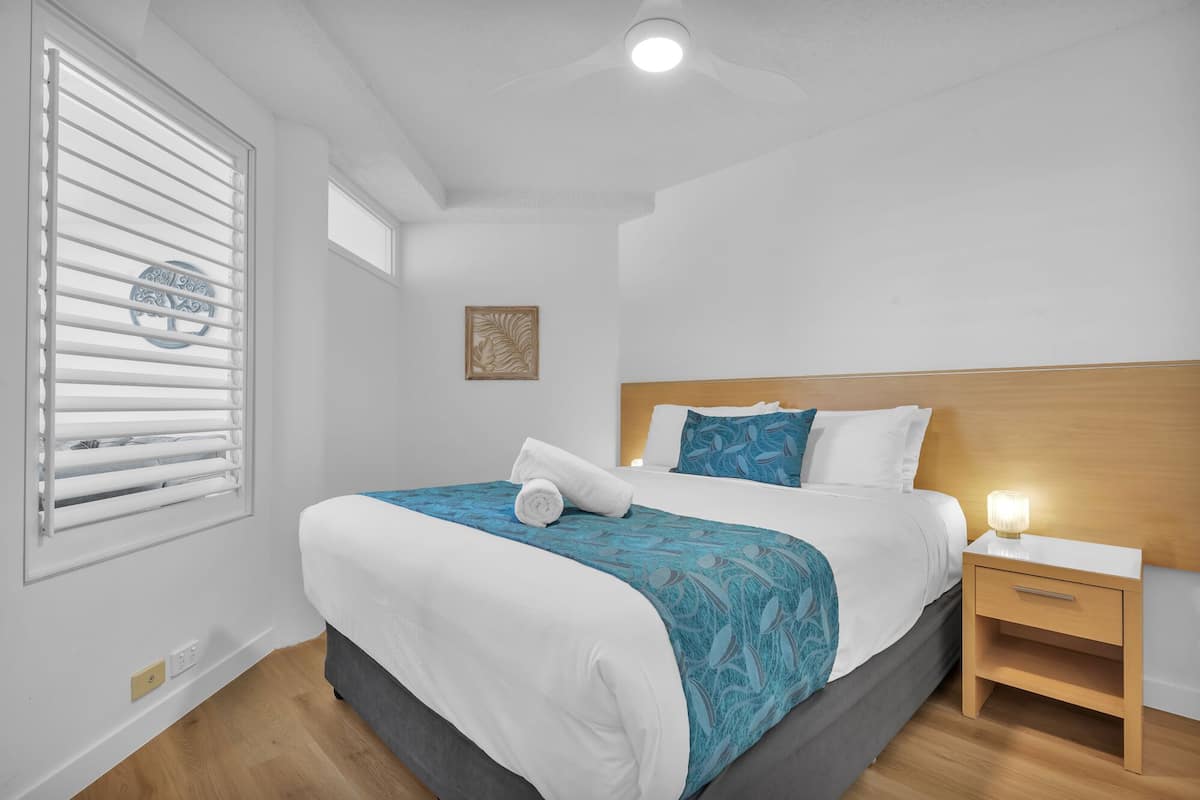 A bedroom with a bed, bedside table, nightstand, window with blinds, and a ceiling light.