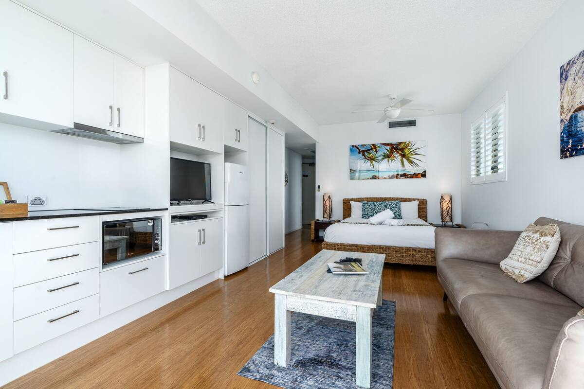 A modern kitchen with white cabinets and a built-in oven. There is a dining area with a wooden table and a sofa. A painting of palm trees is on the wall.