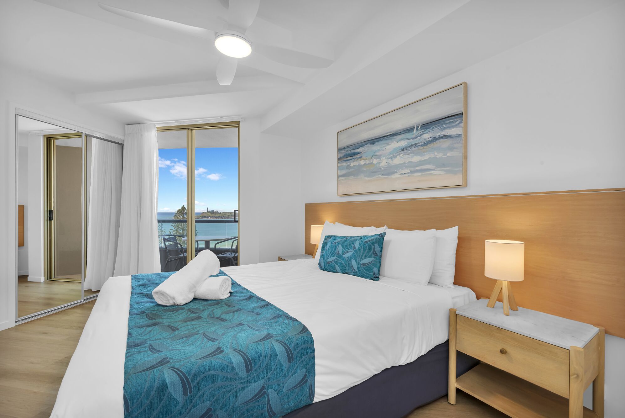 A hotel room with a bed, a nightstand, a painting, and a view of the sea.