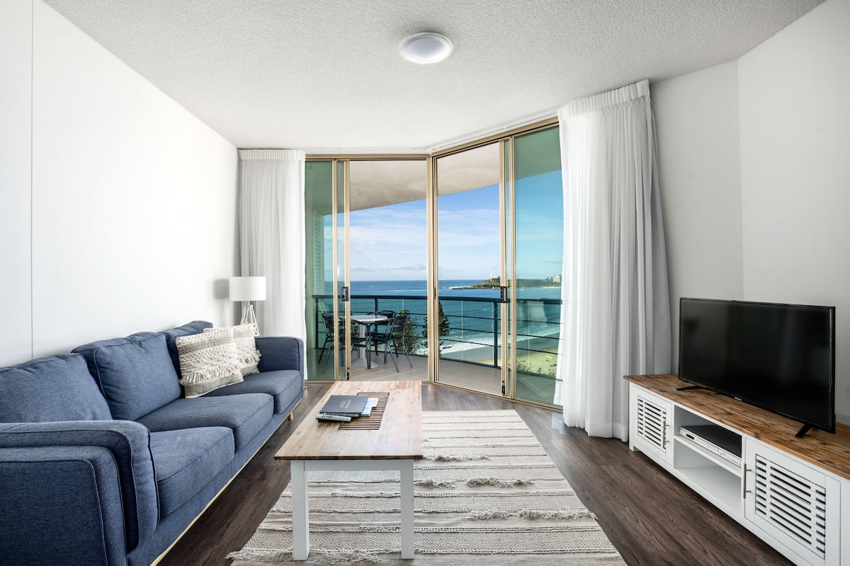 A modern living room with a blue sofa, a wooden coffee table, a flat-screen TV, and a glass door leading to a balcony with a view of the sea.