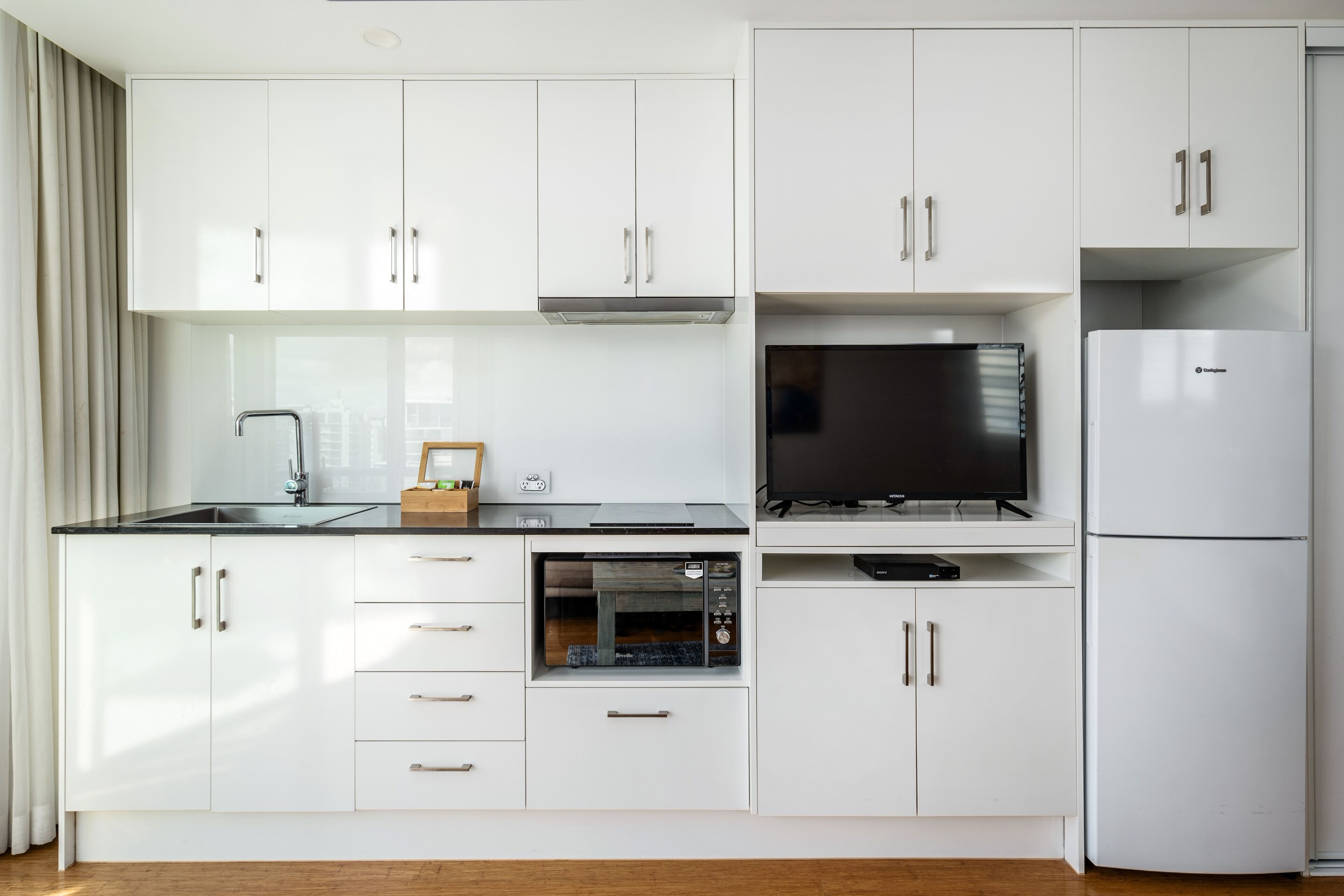Una cocina moderna con armarios blancos, horno integrado, refrigerador y televisor de pantalla plana.