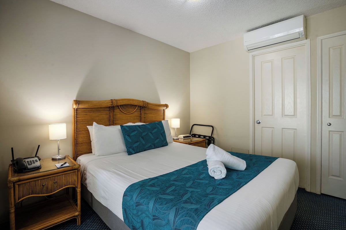 A hotel room with a bed, bedside table with a lamp, a radio, and an air conditioning unit.