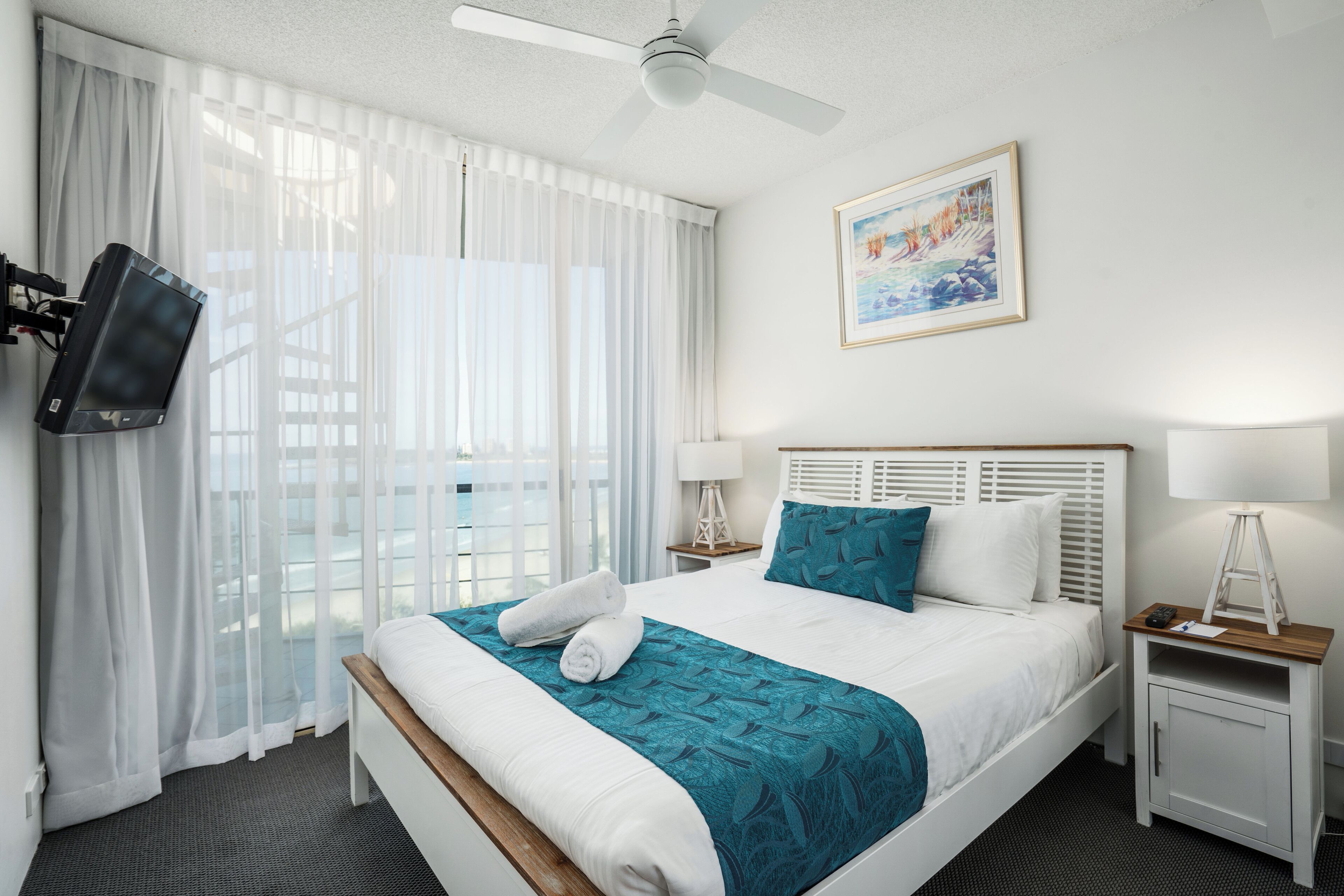 A hotel room with a bed, a television mounted on the wall, a painting, and a window with sheer curtains.