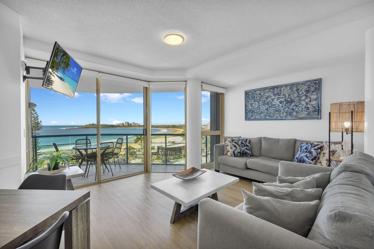 A modern living room with a sofa, coffee table, and a TV. It features large windows with a view of the beach and ocean.
