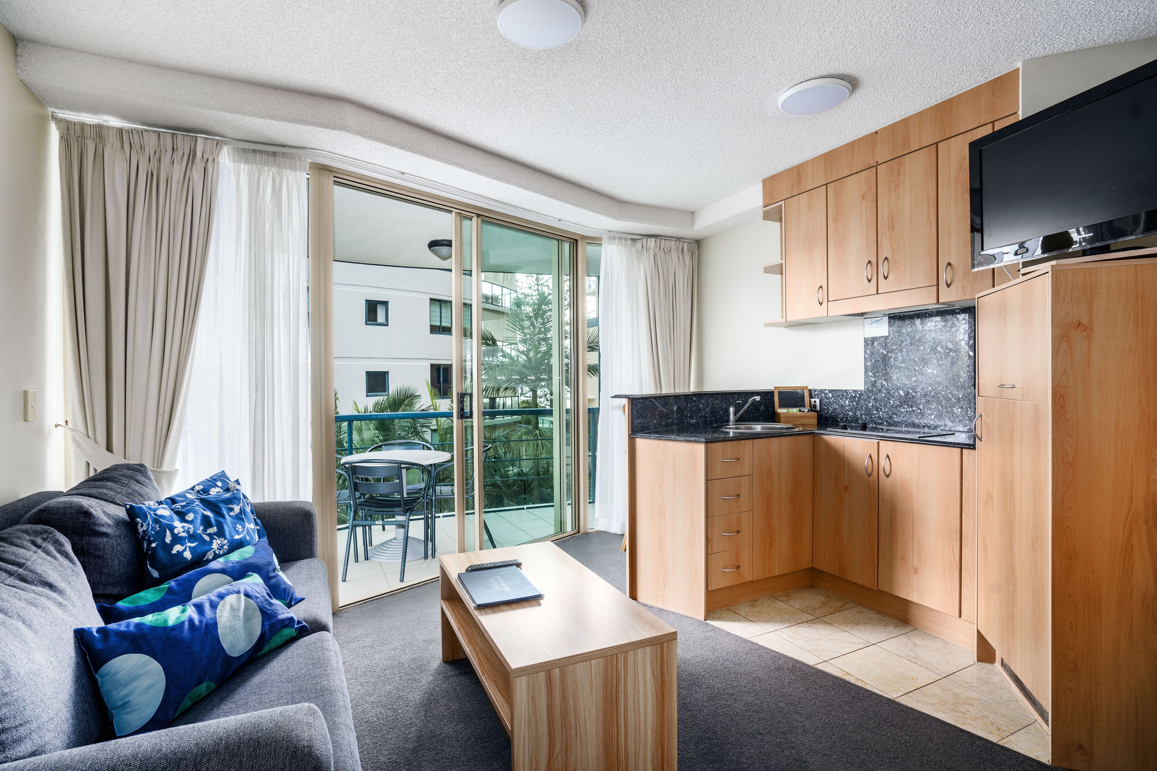 A compact living space with a sofa, a small coffee table, a kitchen area with wooden cabinets, a flat-screen TV, and a sliding glass door leading to an outdoor area.
